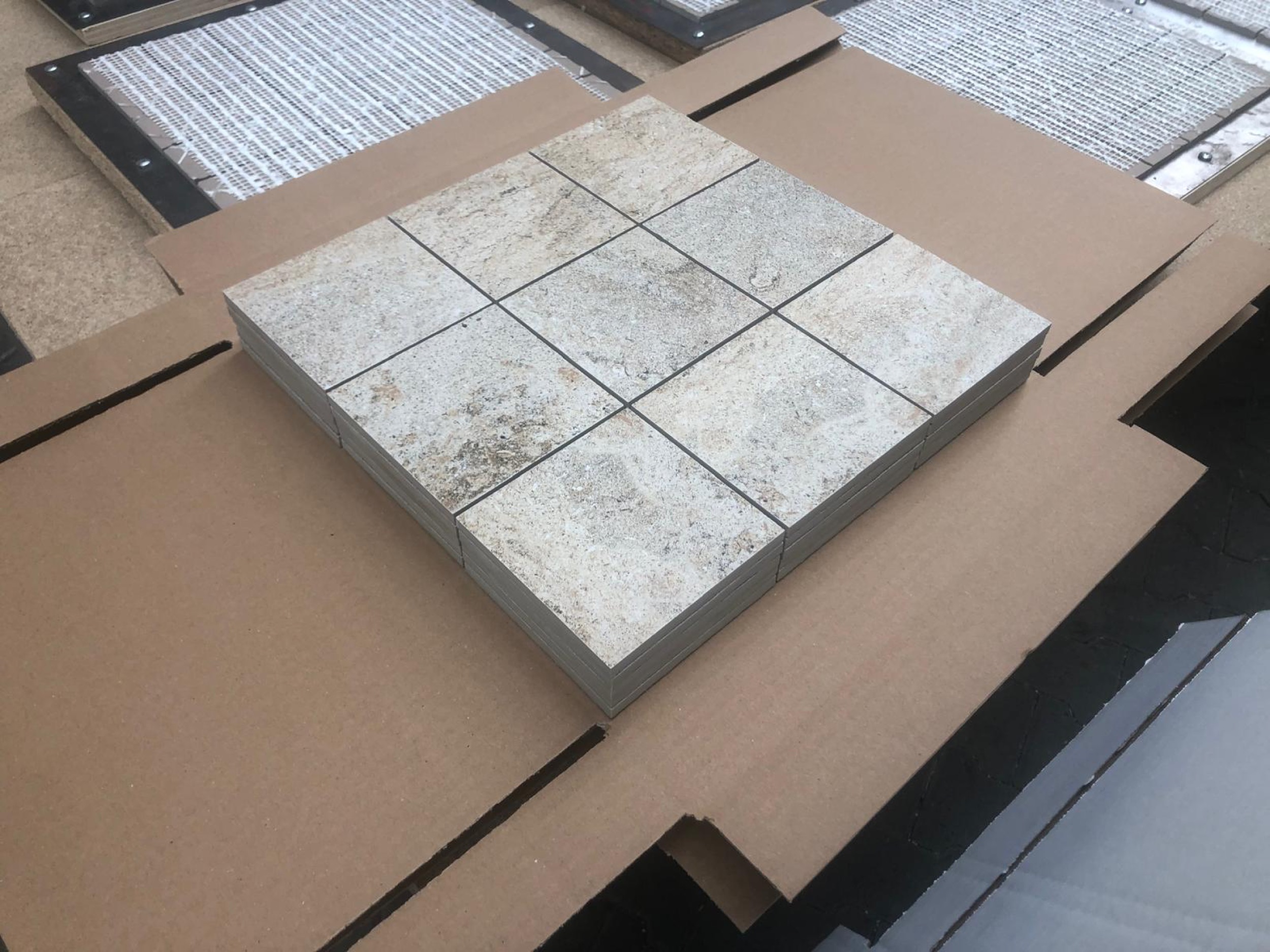 camt neun quadratische steinmosaik platten in beige und grau gestapelt auf geoeffnetem karton in schraeger ansicht